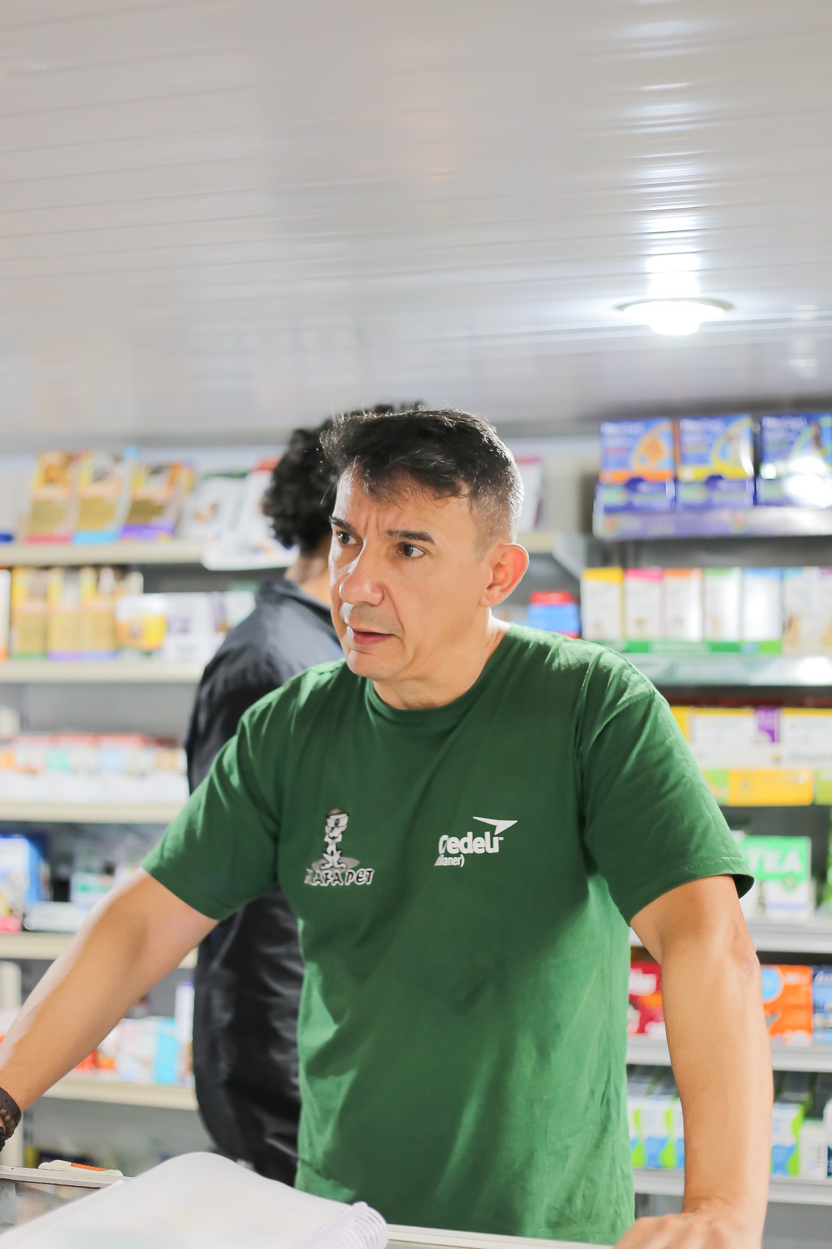 Homem de expressão atenta, vestindo camiseta verde com o logo “Mafa Pet”, está atrás de um balcão em uma loja de produtos veterinários. Ao fundo, prateleiras repletas de medicamentos e itens diversos para pets. A iluminação é clara e o ambiente transmite a rotina de atendimento em um pequeno comércio especializado.