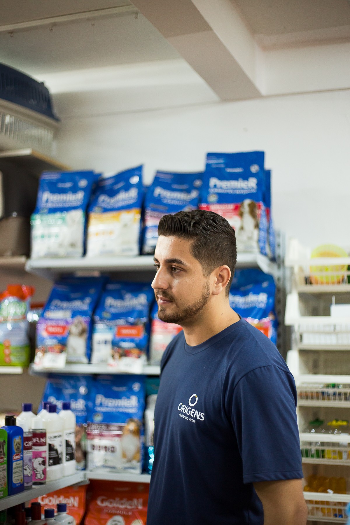 Homem jovem de cabelo curto e barba aparada, usando uma camiseta azul-marinho com o logo “Origens – Nutrindo Amor”. Ele está em uma pet shop, cercado por prateleiras repletas de produtos, incluindo rações das marcas Premier e Golden. O ambiente é bem iluminado e organizado.